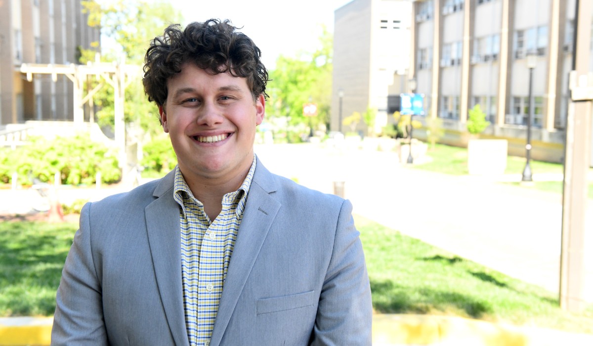 Elijah is sitting on Catholic University's campus. He is wearing a great suit jacket and a checkered shirt.