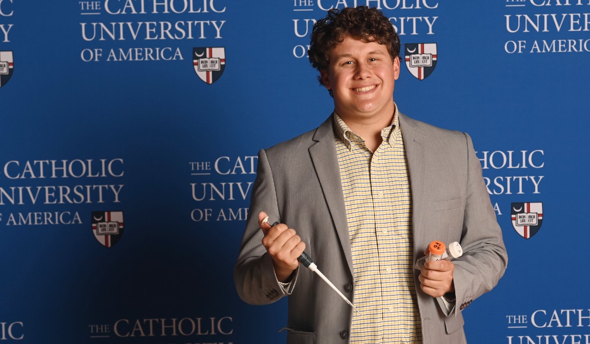 Elijah is standing in front of a Catholic University background. He has curly hair and is wearing a checkered shirt and a grey blazer.