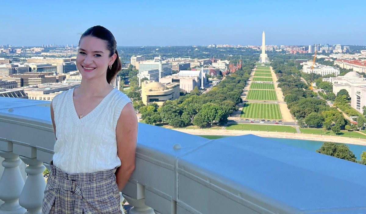 Julia Englert is standing at the Capitol's balcony, looking over the National Mall. She is wearing a white shirt and plaid pants. 