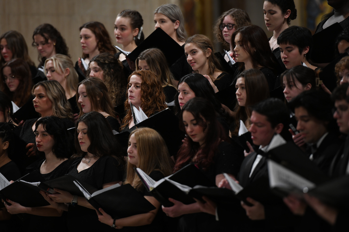 Catholic University Singers look to their conductor as they perform for a crowd. They are all holding sheet music in black binders.