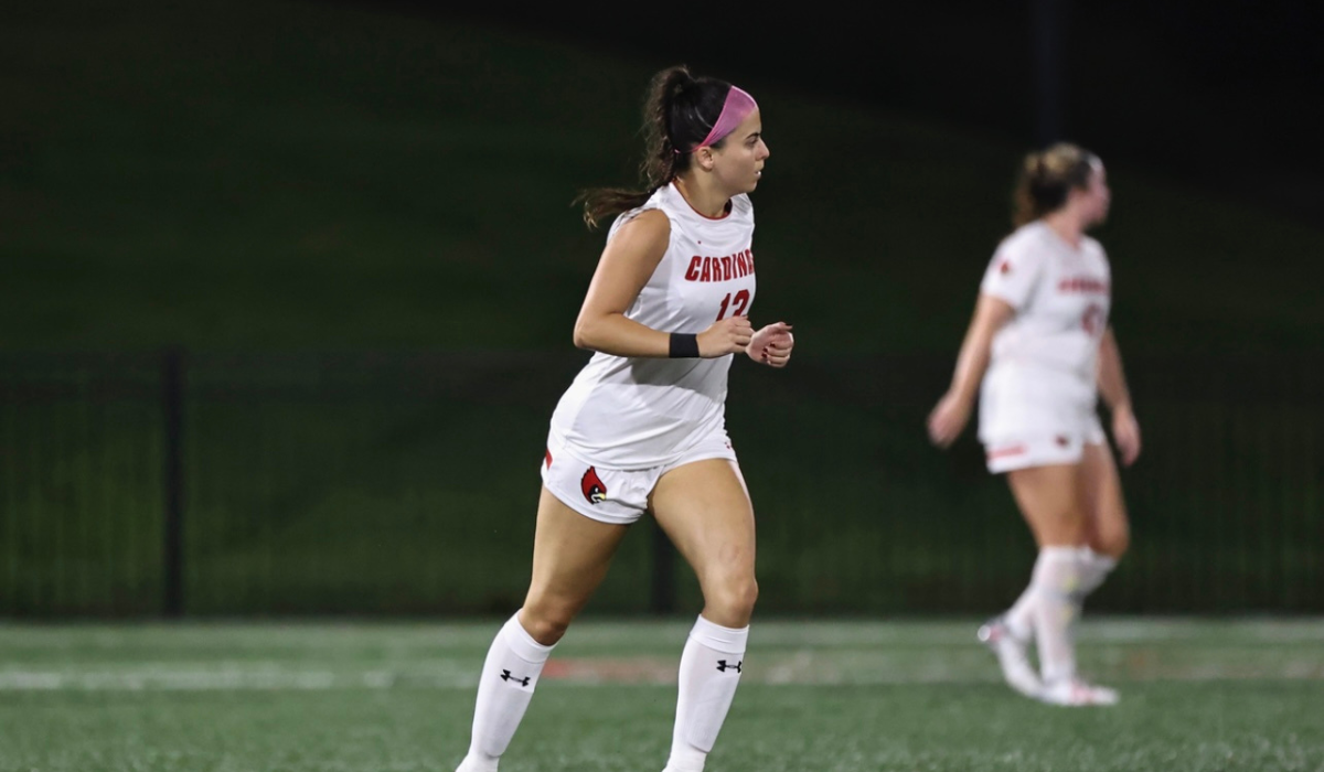 Isabella is on the soccer field at a Catholic University game. She is wearing a white uniform.