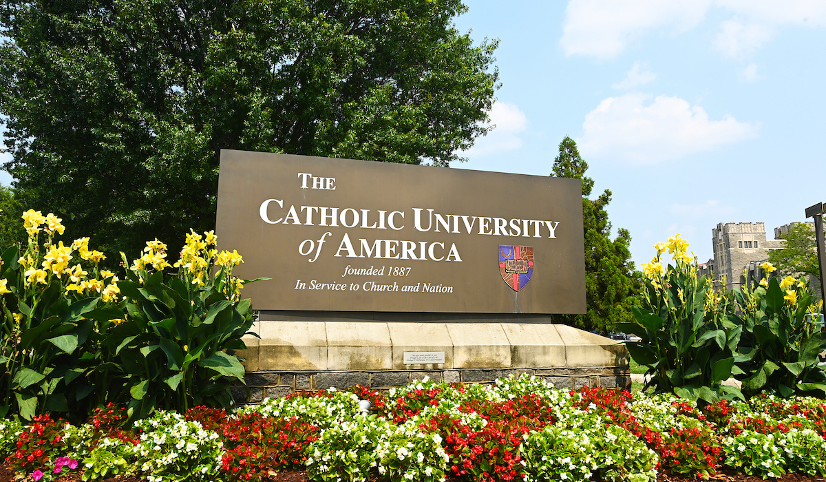 The Catholic University of America sign in summer, surrounded by flowers on a sunny day.