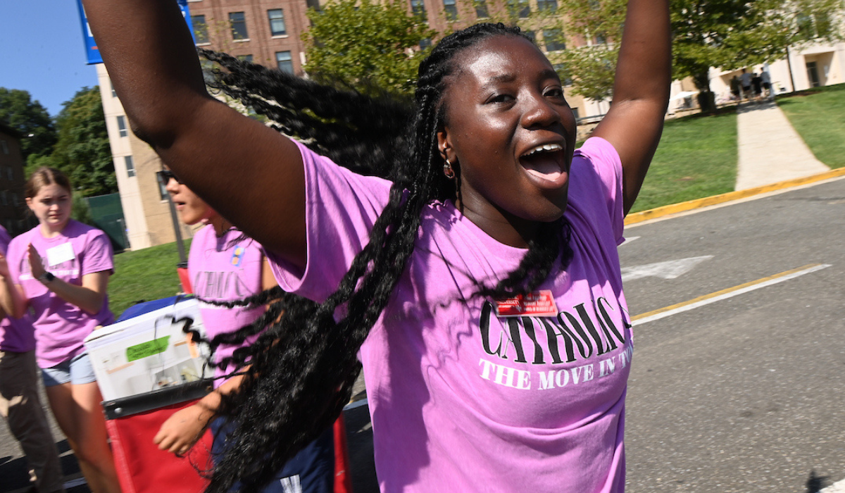 An Orientation Leader throws her arms up in excitement during Move In Day. Behind here, there are two other young women packing a cart to help a new Cardinal settle into their dorm at Catholic University.