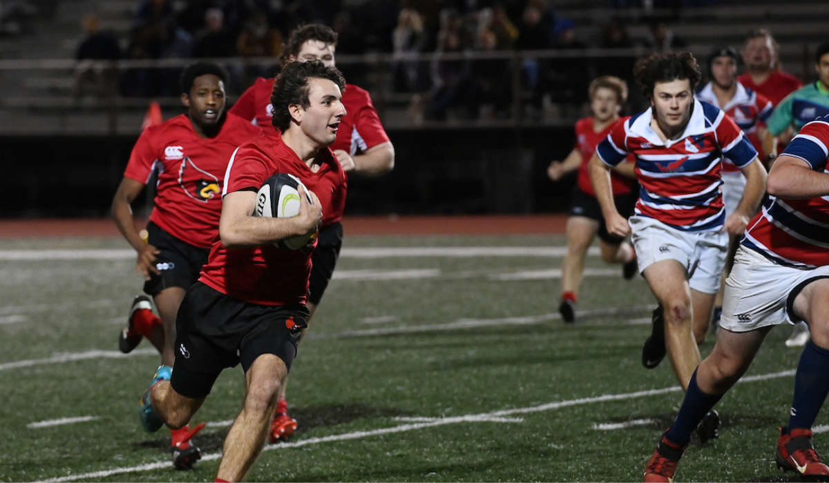 A member of the men's rugby team is holding the ball during a game and running from the pursuit of the other team.