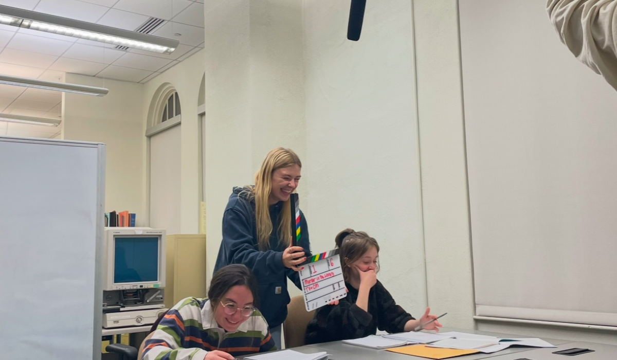 Catherine is directing a scene for her short film, holding a clapboard for a new take between two actors at desks. There is a boom mic overhead.