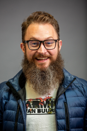 Taylor Black is a man with a long brown and grey beard. He has black, square glasses. He is wearing a grey shirt and a blue puffy vest.