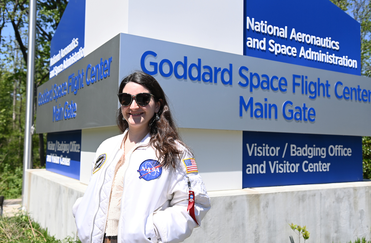 Melissa Kane is standing outside the Goddard Space Flight Center sign. She is wearing a white NASA coat.