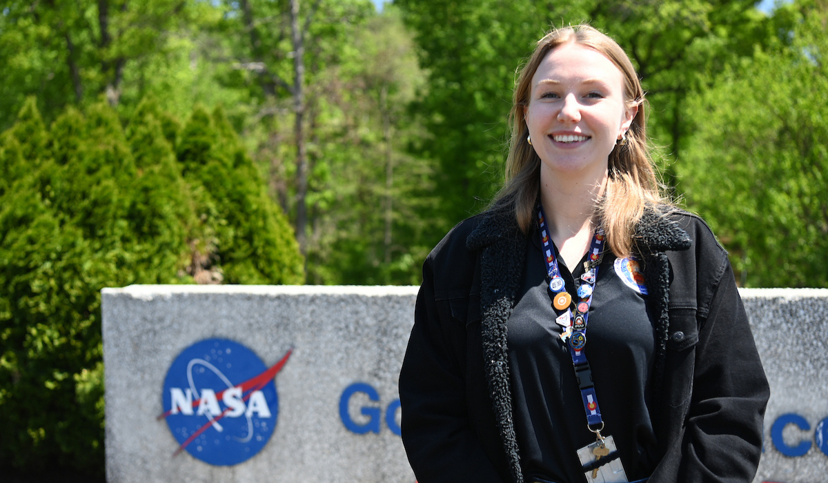 Melissa Kane is standing outside the Goddard Space Flight Center sign. She is wearing a white NASA coat.
