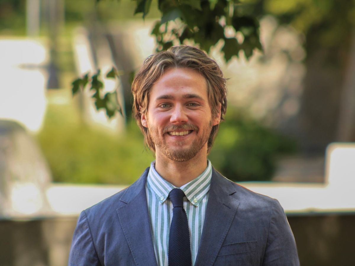 Photo of Nicholas Reynolds. He is outside, surrounded by green trees. He is wearing a blue and white stripe shirt, a navy tie, and a grey suit jacket.