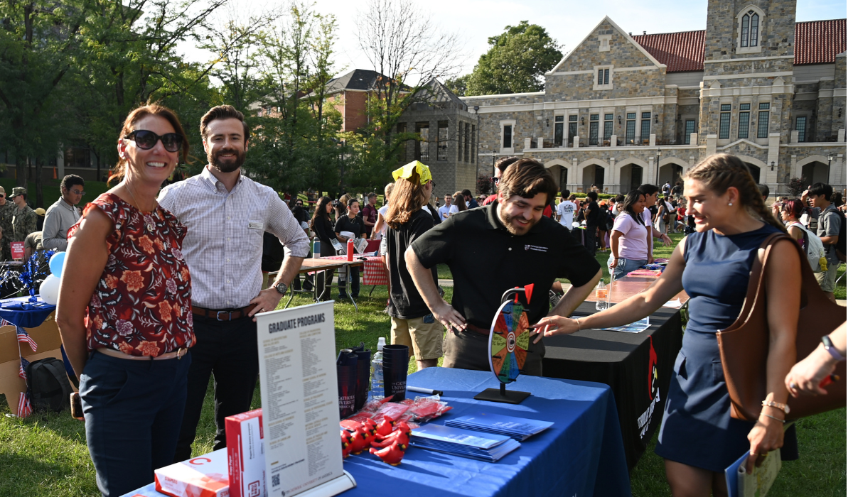Two staff members and two students smile at a Graduate Programs table featuring a prize wheel, informational brochures, and branded merchandise on the University Lawn during Fall Fest.