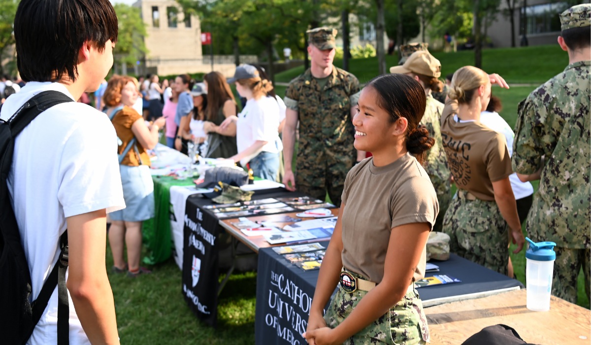A student in military fatigues smiles while talking with a passerby at an ROTC table, with several uniformed service members standing nearby on the University Lawn during Fall Fest.