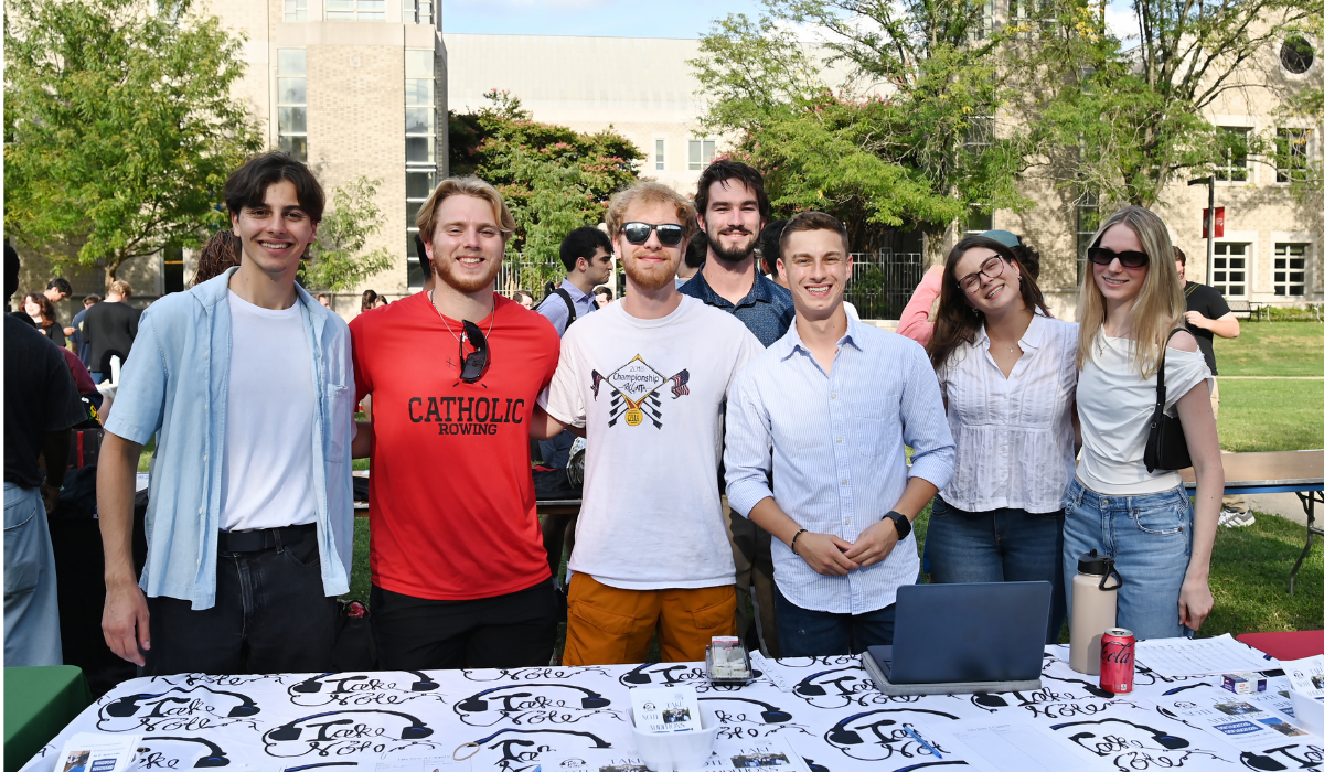 Seven members of a rowing club, one wearing a Catholic Rowing shirt, pose together behind their table during Fall Fest on the University Lawn.