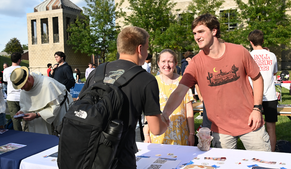 A student shakes hands with a Cardinals for Life club member at an outdoor table while another staff member looks on, during Fall Fest on the University Lawn.