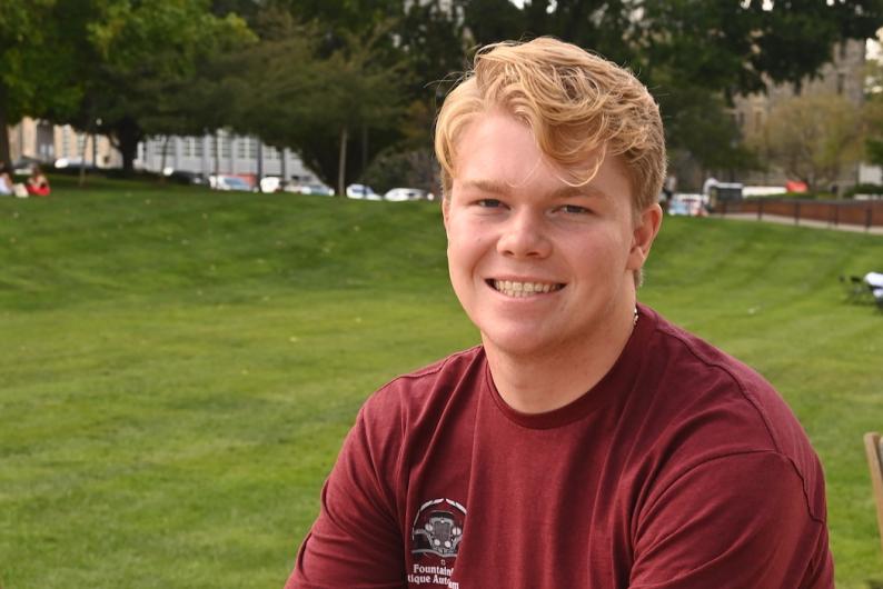 A man with blonde hair and a wide smile, wearing a red shirt, sits in front of a lush green lawn and trees.