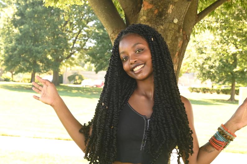 A woman in a black tank top and outstretched arms is standing in front of a tree