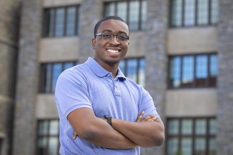 A man stands in front of a grey brick building. He is wearing a blue shirt and has glasses.