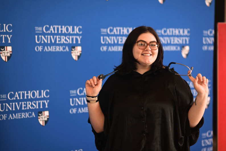 Olivia is standing in front of a blue Catholic University background. She has a stethoscope around her neck.