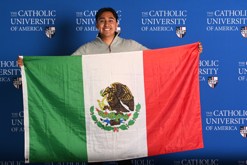 Luis Garcia Abundis stands in front of a blue Catholic University background while holding the Mexican flag.