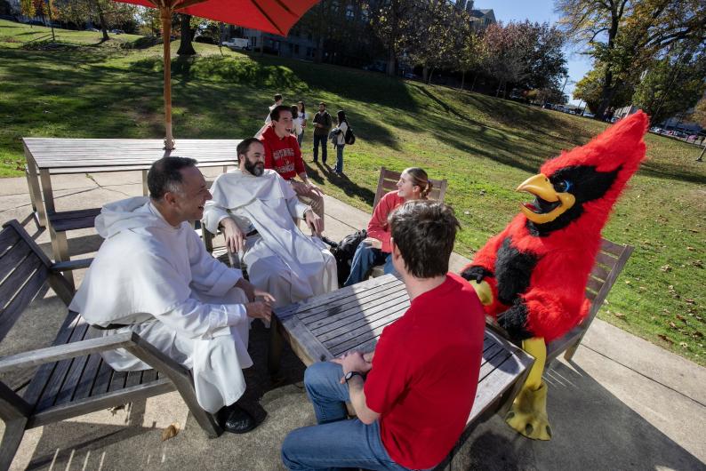 Red, Fr. Aquinas and students chatting at an outdoor table