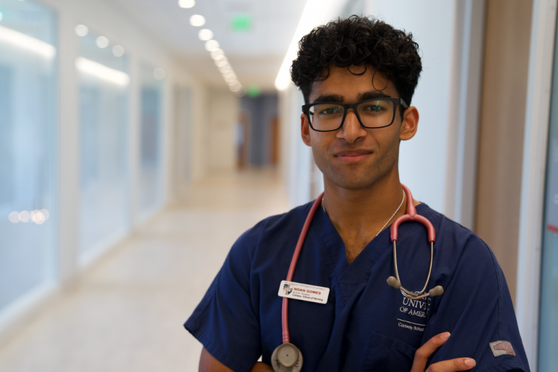 Noah Gomes is standing in the hallway of the Conway School of Nursing. He is wearing blue scrubs and a name tag. A stethoscope is draped around his neck.