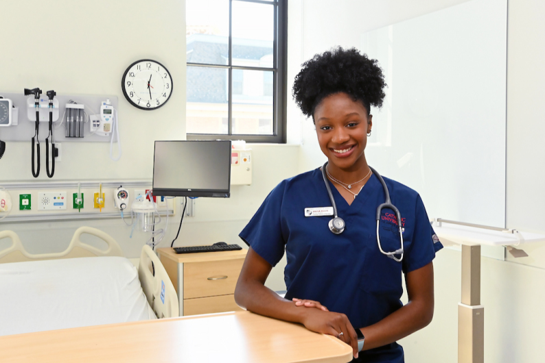 Anisa is in her dark blue scrubs in a hospital room. She has her stethoscope around her neck and is smiling. 
