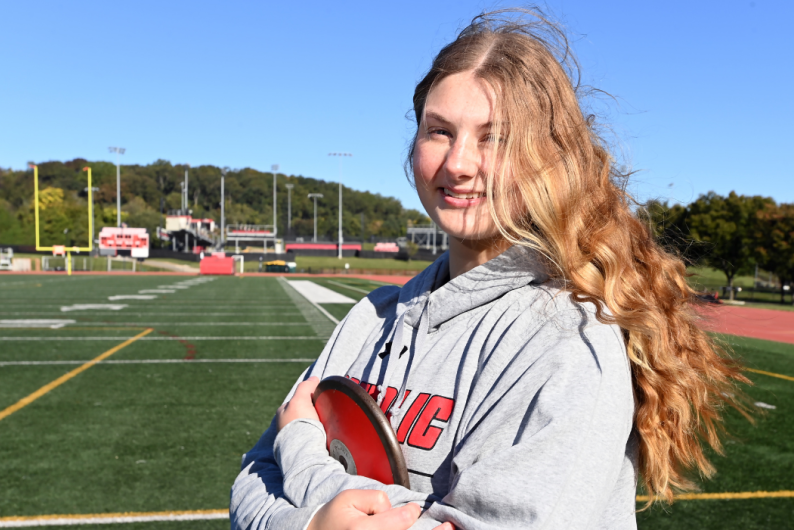 Molly Maxwell is on the track, holding a red discus across her chest. She has long blonde hair and is wearing a grey Catholic University sweatshirt.