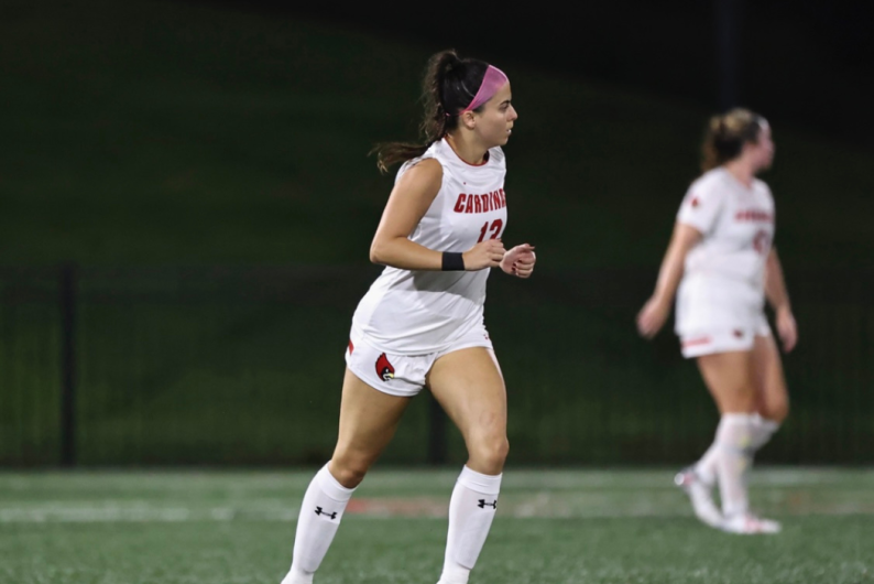 Isabella is on the soccer field at a Catholic University game. She is wearing a white uniform.