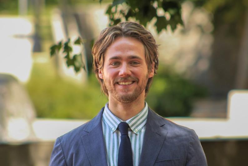 Photo of Nicholas Reynolds. He is outside, surrounded by green trees. He is wearing a blue and white stripe shirt, a navy tie, and a grey suit jacket.